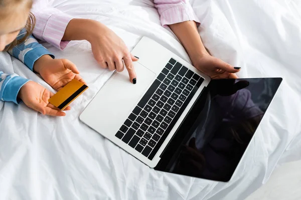 Cropped view of mother using laptop and daughter holding credit card in bedroom — Stock Photo