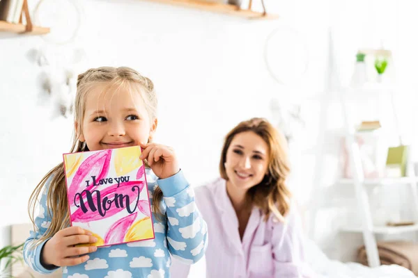 Enfoque selectivo de la hija de la celebración de la tarjeta con te amo mamá letras y sonriente madre en el fondo - foto de stock