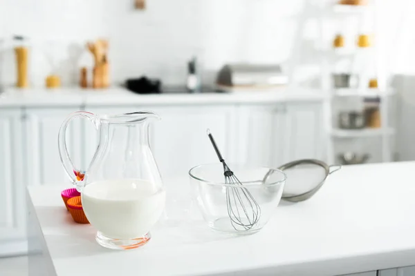 Brocca con latte, ciotola, frusta palloncino, setaccio sul tavolo in cucina — Foto stock