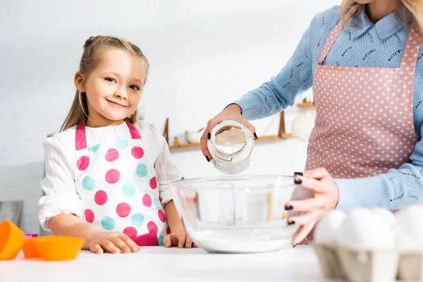 Vue recadrée de mère ajoutant de la farine au bol et fille souriante regardant la caméra — Photo de stock