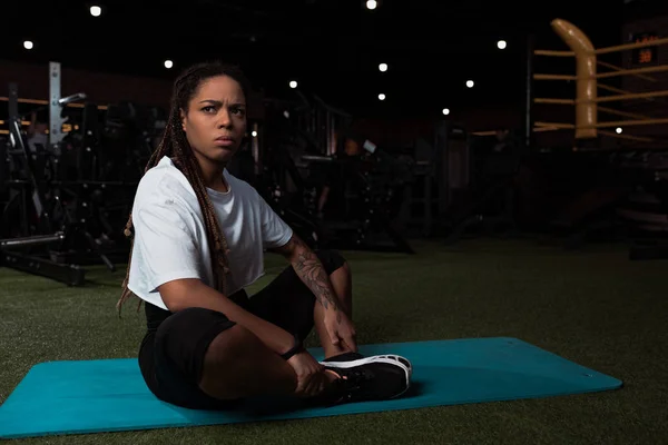 Preoccupata e scontenta donna afroamericana seduta con le gambe incrociate sul tappeto fitness — Foto stock