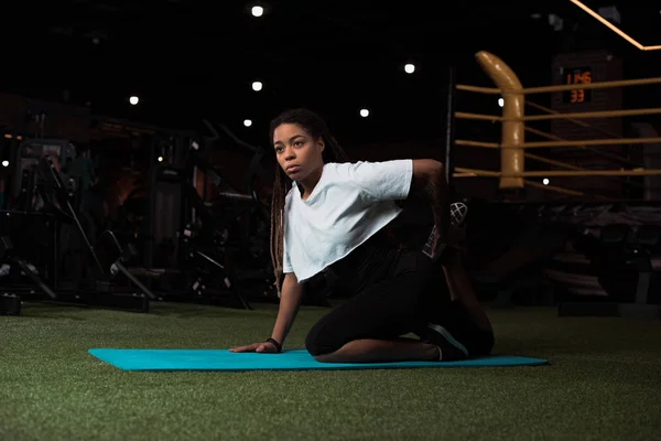 Focus selettivo della donna afroamericana seduta e che si estende sul tappeto fitness — Foto stock