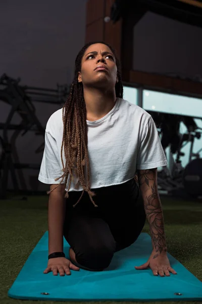 Thoughtful and worried african american woman sitting on fitness mat in gym — Stock Photo