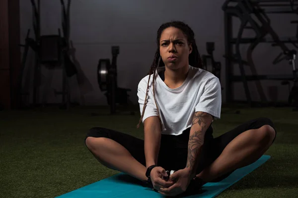 Напряженная африканская американка сидит с сжатыми руками на коврике для фитнеса — стоковое фото