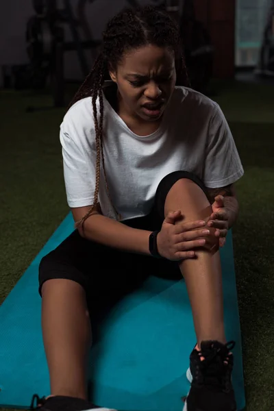 African american woman suffering from pain in leg, sitting on fitness mat — Stock Photo