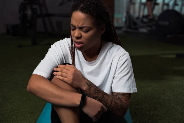 Mujer afroamericana preocupada y molesta que sufre de dolor en la rodilla, sentada en una colchoneta de fitness - foto de stock
