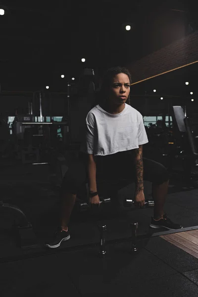 Femme afro-américaine accroupie avec haltères dans la salle de gym — Photo de stock