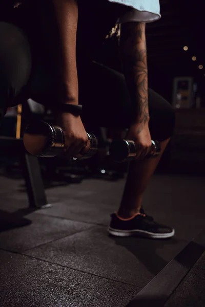 Vista recortada de una mujer afroamericana sentada con pesas en el gimnasio - foto de stock