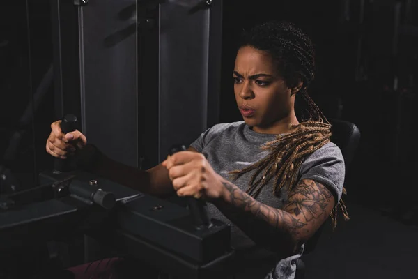 Selective focus of tattooed african american girl with dreadlocks working out on fitness machine — Stock Photo