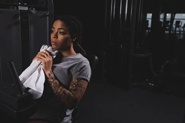 Mujer afroamericana tatuada sosteniendo la toalla cerca de la máquina de fitness - foto de stock