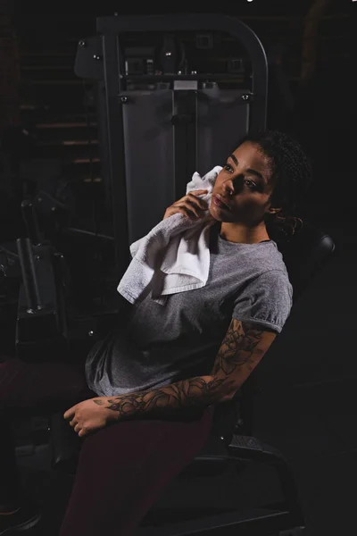 Chica afroamericana tatuada sosteniendo la toalla cerca de la máquina de fitness - foto de stock