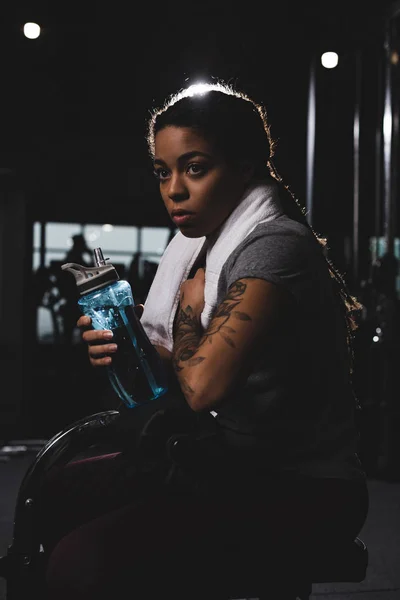 Jeune fille afro-américaine avec tatouage tenant bouteille de sport dans la salle de gym — Photo de stock