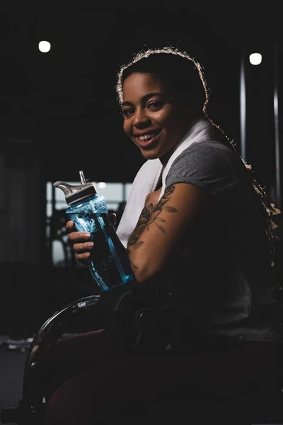 Chica afro-americana feliz con tatuaje sosteniendo botella deportiva en el gimnasio — Stock Photo