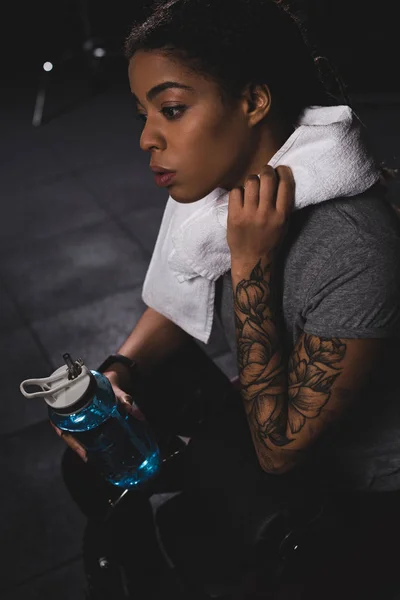 Attraente ragazza afro-americana con tatuaggio in possesso di bottiglia sportiva in palestra — Foto stock