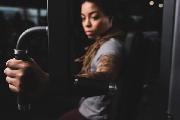 Messa a fuoco selettiva di attraente ragazza afro-americana con tatuaggio esercizio sulla macchina palestra — Foto stock