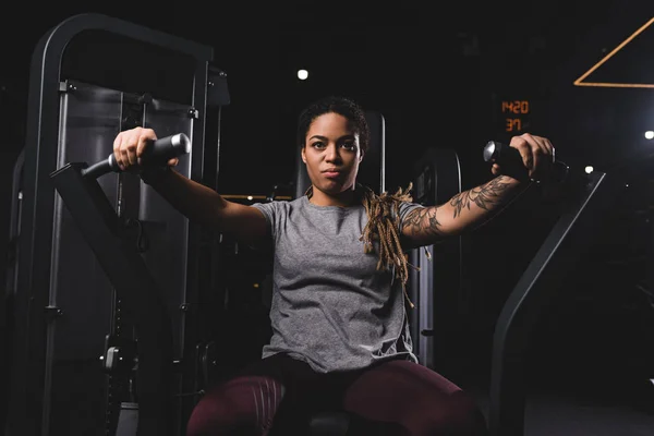 Atlética y tatuada mujer afroamericana haciendo ejercicio en la máquina de gimnasio - foto de stock