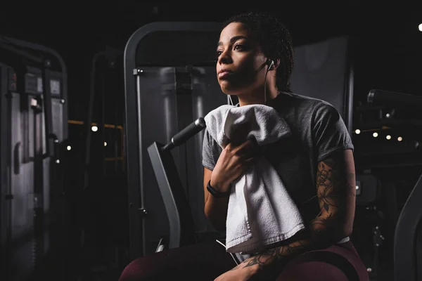 Mujer afroamericana tatuada escuchando música y sosteniendo la toalla en el gimnasio - foto de stock