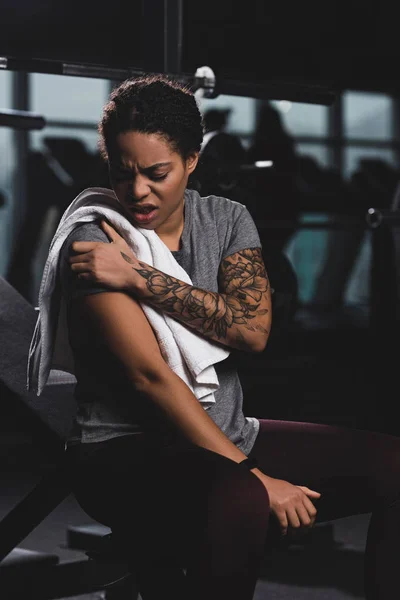 Injured african american girl with tattoo touching hand while suffering from pain in gym — Stock Photo