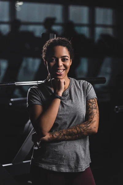 Happy african american girl with tattoo looking at camera in gym — Stock Photo