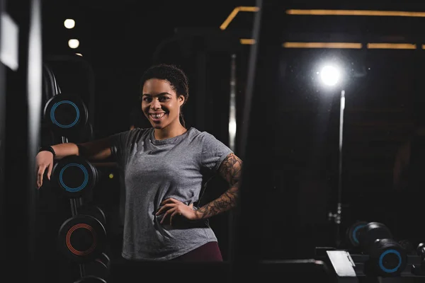 Enfoque selectivo de la chica afroamericana feliz con rastas de pie con la mano en la cadera cerca de las barras en el gimnasio - foto de stock