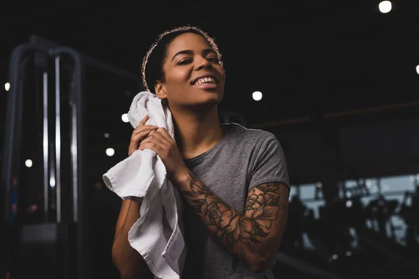 Chica afroamericana tatuada limpiando el sudor con toalla y sonriendo n gimnasio - foto de stock