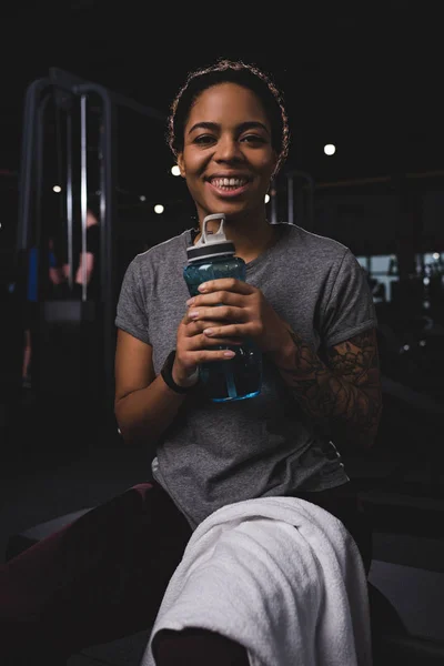 Mujer afroamericana feliz y tatuada sosteniendo botella deportiva - foto de stock