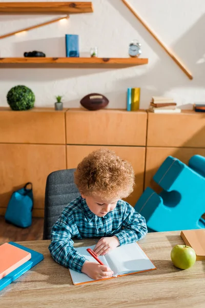 Cute smart kid writing in notebook near books — Stock Photo