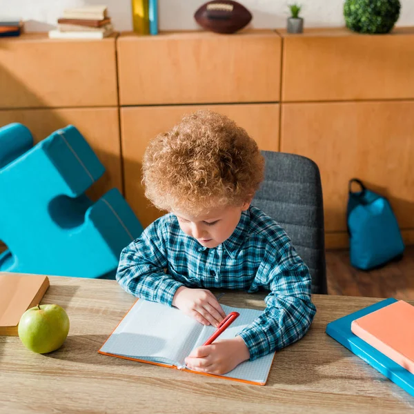 Милий розумний малюк пише в блокноті біля книг і яблука — стокове фото