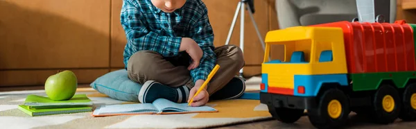 Panoramic shot of smart kid writing in notebook near toy car and apple while sitting on floor — Stock Photo
