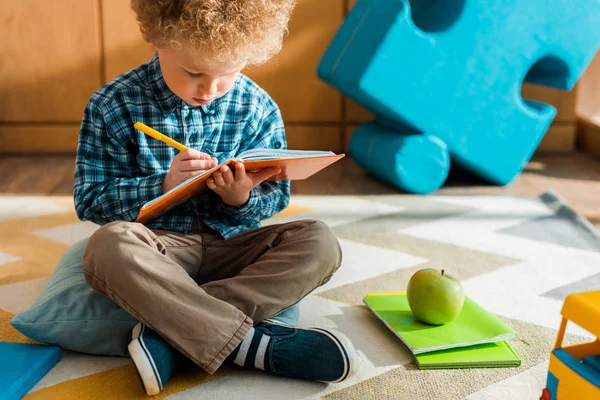 Niño inteligente escribiendo en un cuaderno mientras está sentado en el suelo cerca de la manzana verde - foto de stock