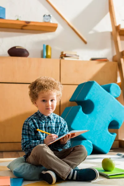 Smart kid écrit dans un cahier tout en étant assis sur le sol près de pomme et livres — Photo de stock