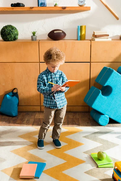 Niño lindo e inteligente escribiendo en el cuaderno, mientras que de pie cerca de libros y manzana - foto de stock