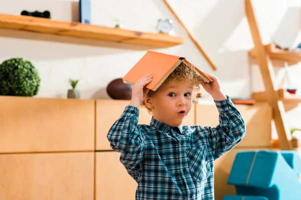 Sorprendido e inteligente niño sosteniendo libro por encima de la cabeza en casa - foto de stock