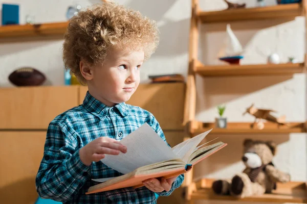 Bambino riccio che tiene il libro e guarda lontano a casa — Foto stock