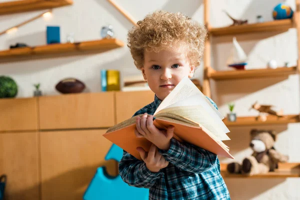 Niño rizado sosteniendo libro y mirando a la cámara en casa - foto de stock