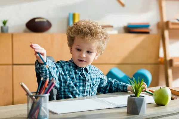 Selective focus of smart and curly kid taking pencil near ripe apple on table — Stock Photo