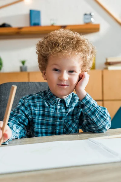 Избирательный фокус счастливого и кудрявого ребенка, держащего карандаш рядом с чистой бумагой — стоковое фото