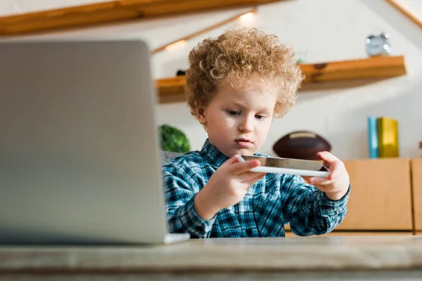 Messa a fuoco selettiva di bambino intelligente guardando smartphone vicino al computer portatile — Foto stock