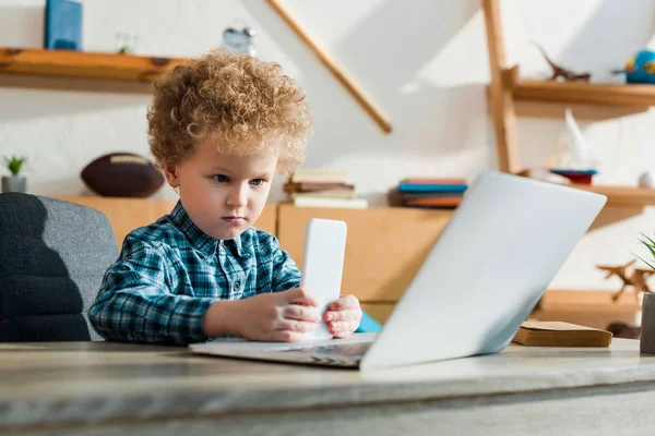 Enfoque selectivo de inteligente niño sosteniendo teléfono inteligente cerca de la computadora portátil - foto de stock