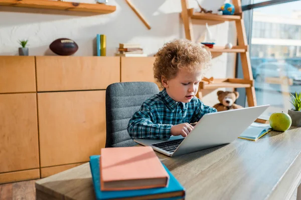 Селективное внимание кудрявого ребенка, печатающего на ноутбуке рядом с книгами — стоковое фото