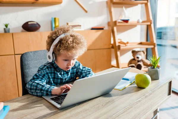 Кудрявый ребенок печатает на ноутбуке рядом с яблоком, слушая музыку в беспроводных наушниках — стоковое фото