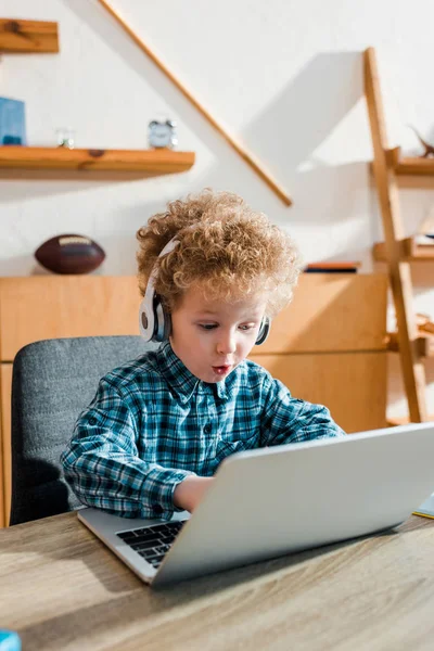 Selektiver Fokus des überraschten Kindes, das während des Musikhörens in drahtlosen Kopfhörern auf dem Laptop tippt — Stockfoto