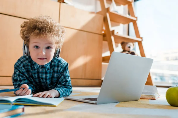 Селективное внимание умного ребенка, слушающего музыку в беспроводных наушниках, держа ручку рядом с ноутбуком — стоковое фото