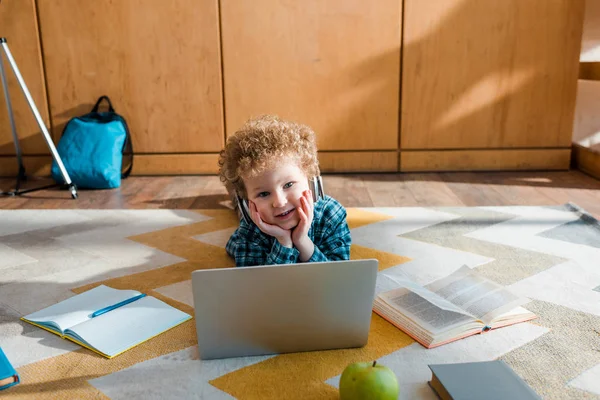 Bambino felice con le cuffie sdraiato sul pavimento vicino al computer portatile — Foto stock