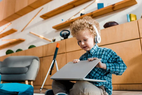 Garoto bonito usando laptop enquanto ouve música em fones de ouvido sem fio — Fotografia de Stock