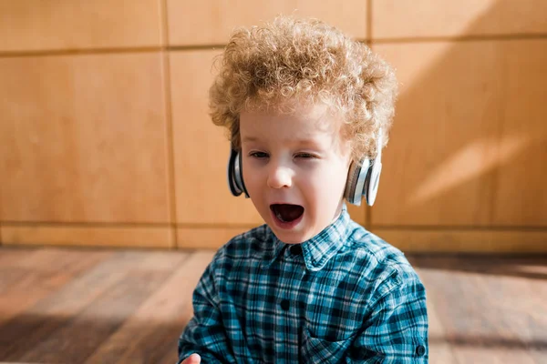 Niño agotado escuchando música en auriculares inalámbricos y bostezando en casa - foto de stock