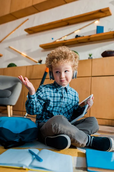 Selective focus of confused kid in headphones holding digital tablet and showing shrug gesture — Stock Photo