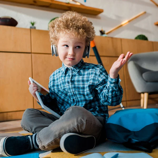 Selective focus of confused kid in headphones holding digital tablet and showing shrug gesture — Stock Photo