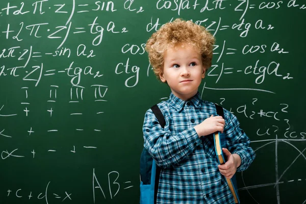 Smart kid standing with book near chalkboard with mathematical formulas — Stock Photo