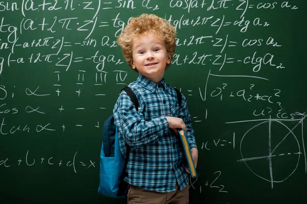 Criança feliz segurando livro perto de quadro-negro com fórmulas matemáticas — Fotografia de Stock